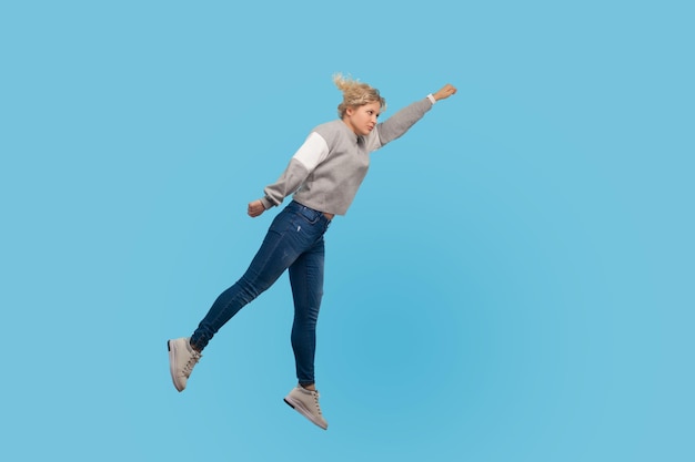 Superman Ritratto a figura intera di una donna determinata in felpa e jeans che vola in aria sentendosi superpotere e libertà trampolino per il successo in studio indoor girato isolato su sfondo blu