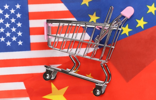 Supermercato internazionale e globale. Mini carrello della spesa sullo sfondo della bandiera sfocata degli Stati Uniti, della Cina e dell'Unione europea. Concetto di acquisto.