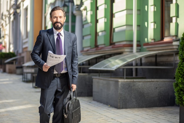 Supporti stampati. Uomo d'affari bello positivo che cammina per lavorare mentre si tiene un quotidiano