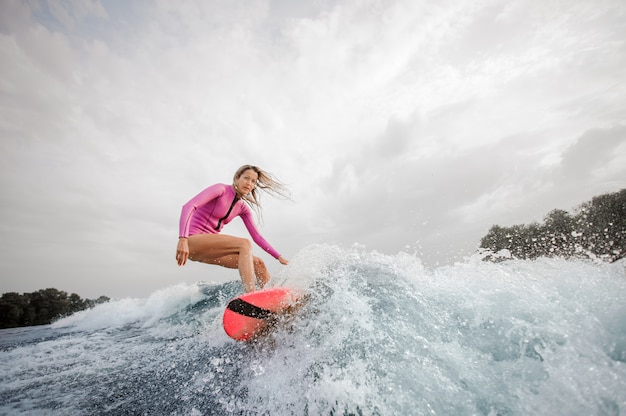 Surfista biondo della donna che guida giù l'onda di spruzzatura blu contro il cielo