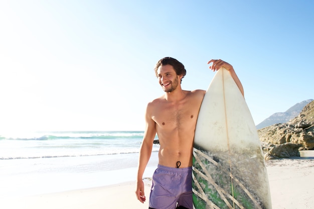 Surfista maschio felice che sta con il suo bordo alla spiaggia