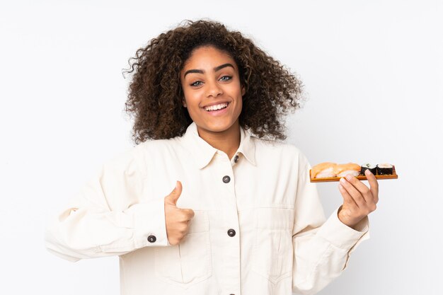 Sushi della giovane donna afroamericana della tenuta sulla parete bianca con i pollici su perché è successo qualcosa di buono