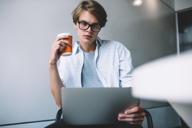 sviluppatore di software maschile con bicchiere di carta seduto in uno spazio di coworking con internet ad alta velocità sul laptop