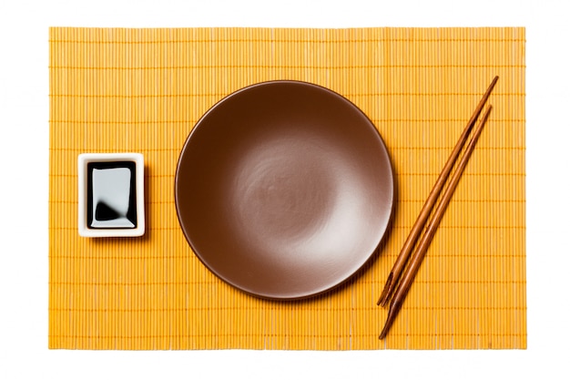 Svuoti il piatto marrone rotondo con le bacchette per i sushi e la salsa di soia sulla stuoia di bambù gialla