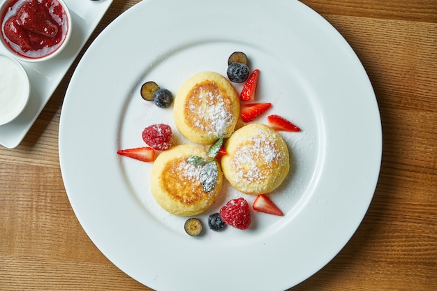 Syrniki ucraino classico con frutti di bosco, marmellata e panna acida in un piatto bianco su un tavolo di legno. Dessert appetitoso per la colazione