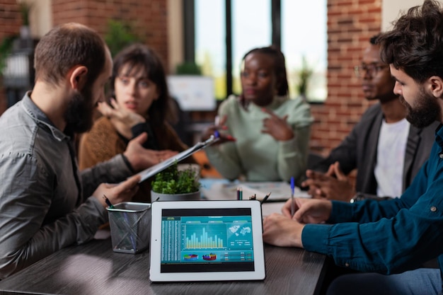 Tablet computer con fatturato aziendale sullo schermo in piedi sul tavolo nell'ufficio di avvio. Uomini d'affari multietnici millenari che discutono della strategia di marketing che lavorano alla collaborazione aziendale