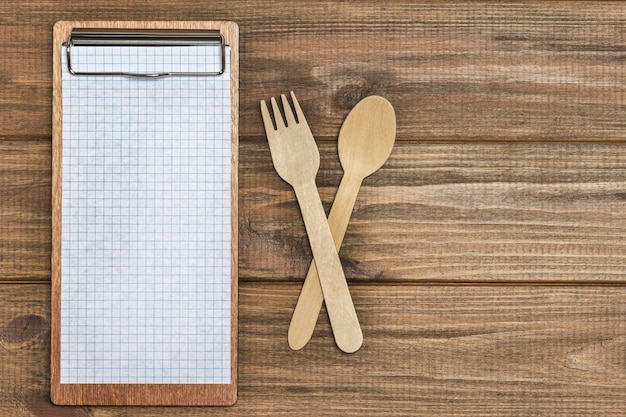 Tablet menu con foglio bianco e forchetta e cucchiaio in legno. Concetto di nutrizione alimentare sana. Copia spazio. Fondo in legno. Disposizione piatta.