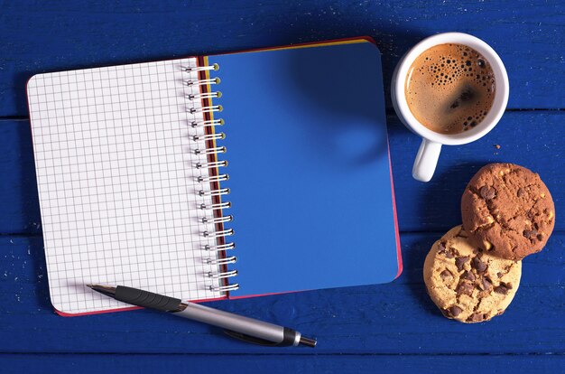Taccuino aperto, tazza di caffè e biscotti su uno sfondo blu scuro, vista dall'alto