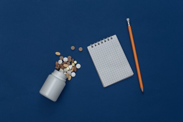 Taccuino con matita, bottiglia di pillole su sfondo blu classico. Colore 2020. Vista dall'alto