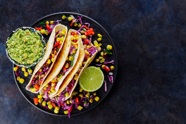 Tacos vegetariani con mais dolce, cavolo viola e pomodori su un piatto nero. Tacos con verdure e salsa guacamole su sfondo scuro. Copia spazio. Vista dall'alto