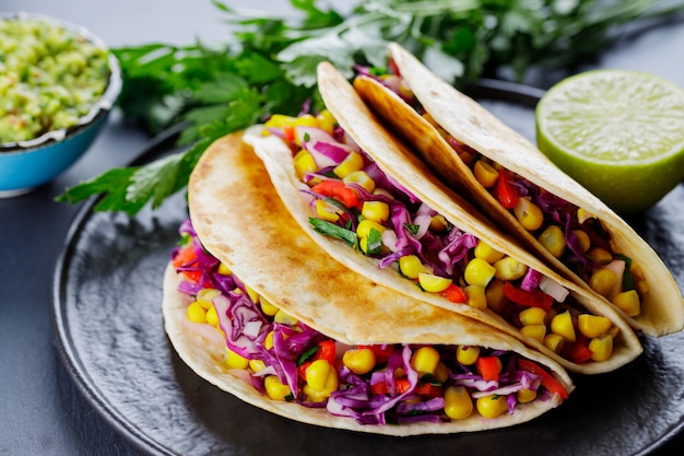 Tacos vegetariani con varie verdure, guacamole e lime su sfondo scuro. Tacos con mais dolce, cavolo viola e pomodori su un piatto nero. Avvicinamento