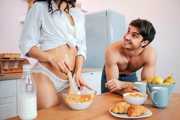 Tagli il punto di vista del supporto sexy della giovane donna in cucina alla tavola. Mescola i fiocchi di mais con il latte in una ciotola. Eccitato giovane guardarla e sorridere. Tiene la tazza.