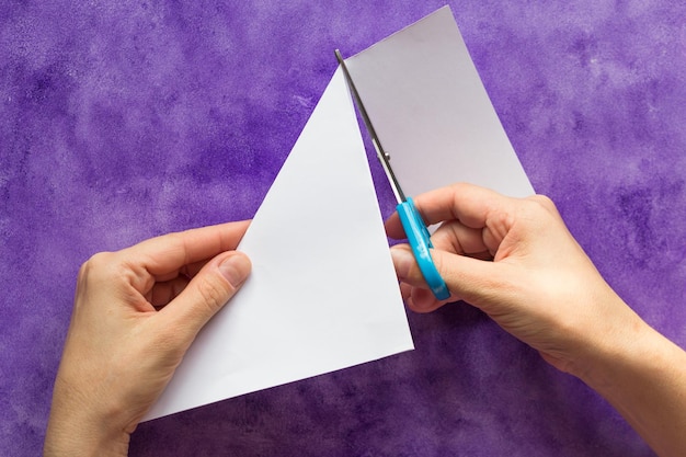 Tagliare il bordo del foglio di carta con l'angolo piegato delle forbici per fare un pezzo quadrato dalle mani della donna sulla superficie viola