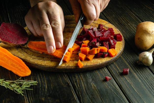 Tagliare le carote e le barbabietole bollite in piccoli pezzi per preparare la vinaigrette Un coltello in mano a un cuoco sul tavolo della cucina Concetto di piatto di verdure su sfondo scuro