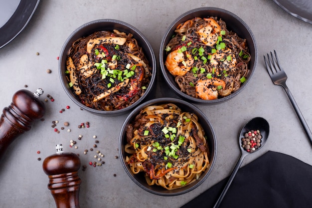 Tagliatelle al wok con gamberi, in un piatto nero decorato con piselli, su uno sfondo grigio. Il concetto di cucina asiatica.