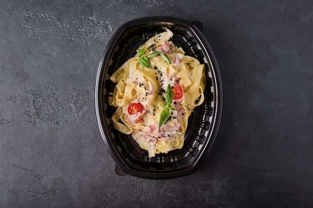 Tagliatelle alla carbonara in contenitore di plastica nera su sfondo grafite scuro vista dall'alto designer food