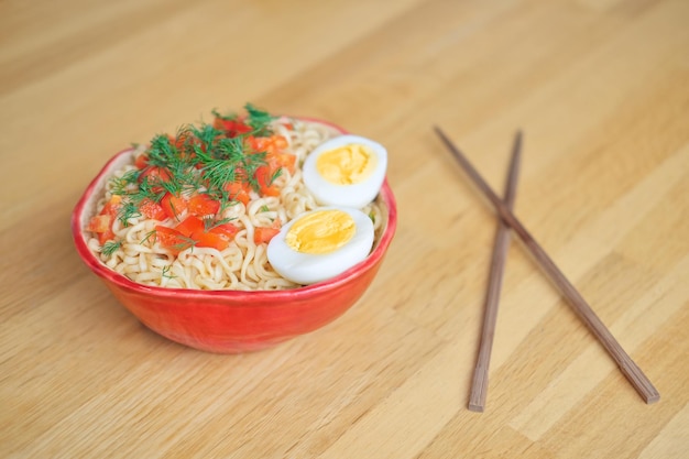 tagliatelle asiatiche con uova e verdure in una ciotola rossa e bacchette cinesi su un tavolo di legno