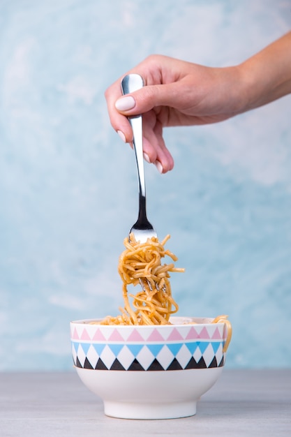 Tagliatelle con salsa in una ciotola colorata, mano tiene una forchetta di pasta appesa, spaghetti appetito sul blu.