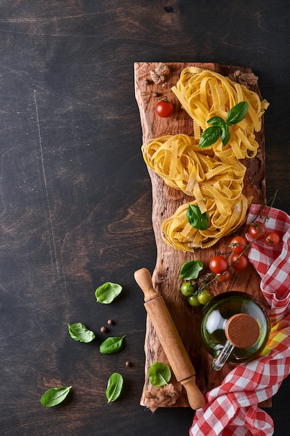 Tagliatelle. Pasta fatta in casa, foglie di basilico, farina, pepe, olio d'oliva, pomodorini e mattarello e coltello da pasta su fondo di legno vecchio scuro. Concetto di cibo. Modello. Orizzontale con copia spazio.