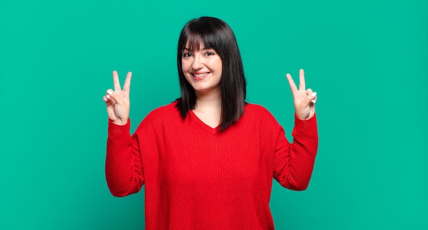 Taglie forti donna graziosa che sorride e sembra felice, amichevole e soddisfatta, gesticolando vittoria o pace con entrambe le mani