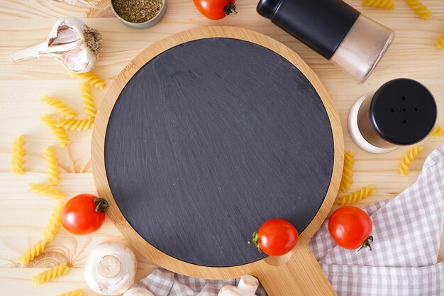 Tagliere di legno e ingredienti freschi per cucinare: pasta, pomodoro e spezie sul tavolo in legno, vista dall'alto.