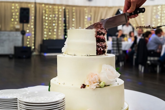 Taglio del primo livello della torta nuziale di mastice bianco sullo sfondo della sala banchetti. Vista frontale al tavolo con una bella torta nuziale e piatti laterali per servire gli ospiti. Dividere la torta nuziale in parti.