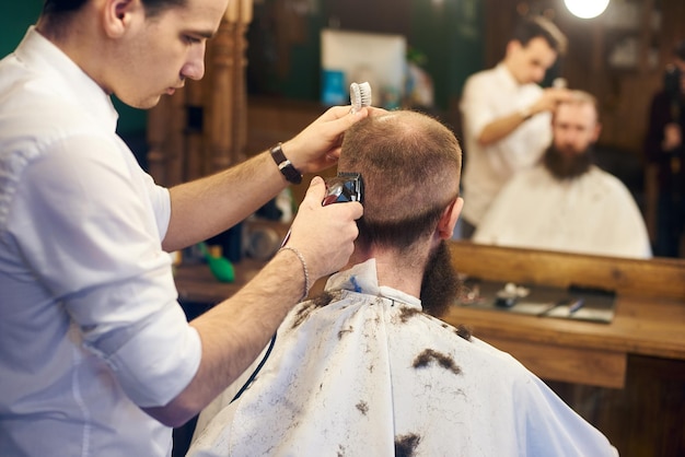 Taglio di capelli corto maschile professionale per cliente barbuto nel negozio di barbiere Giovane barbiere che lavora con un rasoio elettrico Primo piano