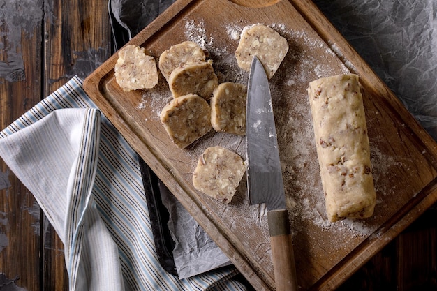 Taglio di pasta frolla fatta in casa per cucinare biscotti di zucchero con noci e cioccolato bianco e coltello da chef su tagliere di legno sul tavolo da cucina in legno scuro. Dolci fatti in casa. Vista dall'alto, spazio