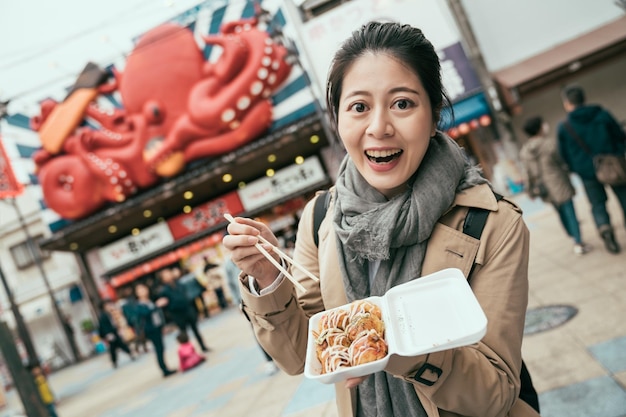 takoyaki in scatola di polistirolo sulle mani di una giovane donna felice eccitante a Osaka in Giappone. viaggiatore coreano che prova il famoso delizioso spuntino giapponese in via tsutenkaku. femmina allegra che mostra cibo locale