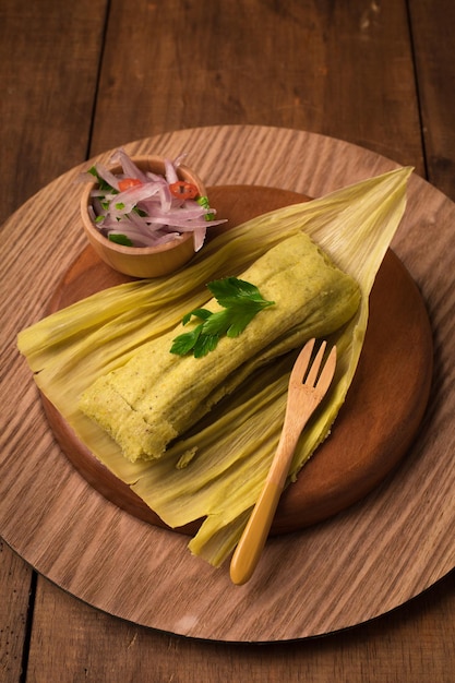 Tamales avvolti in foglie di banano con una ciotola di coriandolo e una forchetta su un piatto di legno.