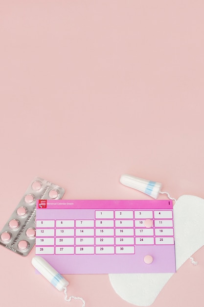 Tamponi, assorbenti femminili e sanitari per giorni critici, calendario femminile, pillole antidolorifiche durante le mestruazioni su una parete rosa. Tracciamento del ciclo mestruale e dell'ovulazione