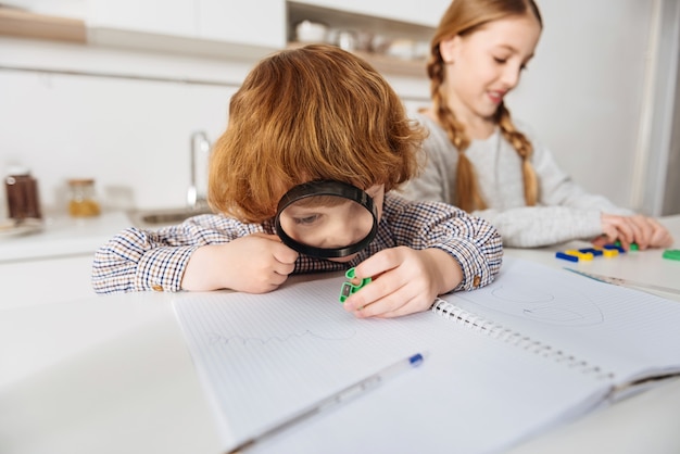 Tanti dettagli. Messo a fuoco vivace ragazzo dello zenzero utilizzando una grande lente guardando uno dei numeri di plastica colorati che sua sorella usando spiegandogli la matematica