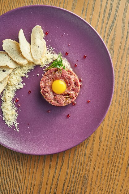 Tartare di manzo fresca con parmigiano, capperi, crostini e quaglia di tuorlo su un piatto viola su una parete di legno