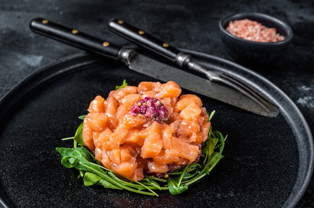 Tartare di salmone crudo o tartare con cipolla rossa, rucola e capperi in banda nera. Sfondo nero. Vista dall'alto.