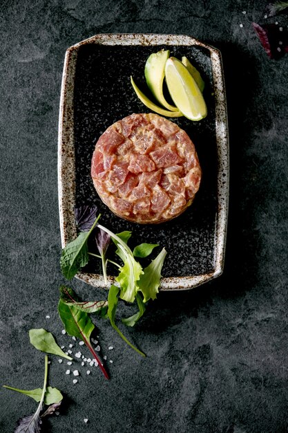 Tartare di tonno con insalata verde, lime, avocado e salsa di senape che serve su piatto in ceramica nera stile giapponese su sfondo di marmo nero. Lay piatto, copia dello spazio. Antipasto al ristorante