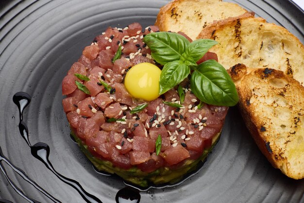 Tartare di tonno e avocado con semi di sesamo, cappri e tuorlo d'uovo su un piatto di ceramica scura con baguette