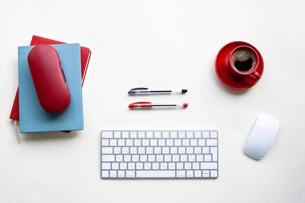 Tastiera e mouse bianchi con penne nere e rosse, tazza di caffè rossa, quaderni sul tavolo bianco