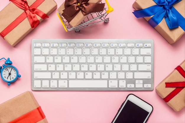 Tastiera, sveglia, cellulare e scatole regalo disposte intorno alla tastiera su una superficie rosa. Concetto di ordinazione e acquisto di regali su Internet, negozio online. Vista piana, vista dall'alto.