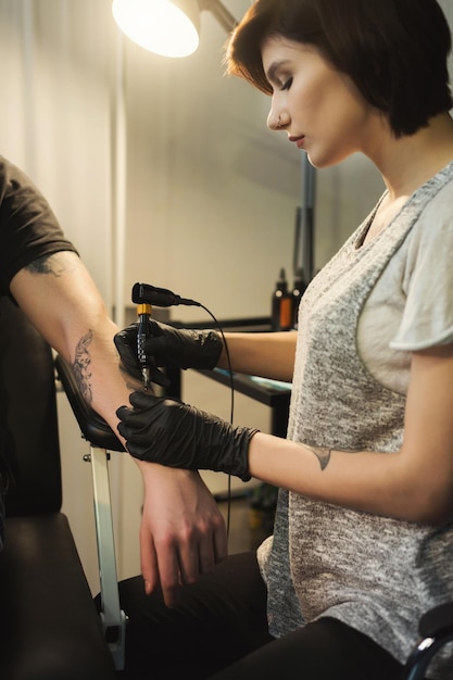 Tatuatore concentrato della donna che fa il tatuaggio con la macchina del tatuaggio sul braccio maschio. Professione femminile insolita, copia spazio