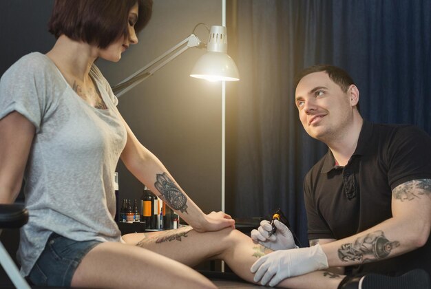 Tatuatore professionista che fa bel tatuaggio sulla gamba della giovane donna. Uomo tatuatore disegno immagine sul corpo femminile con macchina speciale, copia spazio