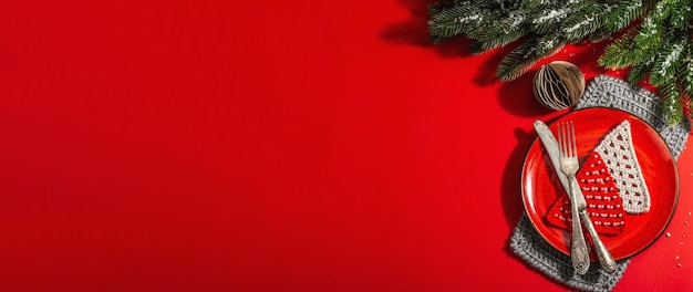 Tavola di Natale servita nei toni del rosso con posate vintage. Rami sempreverdi, abeti lavorati all'uncinetto, neve artificiale. Luce dura moderna, ombra scura. Piatto lay, sfondo rosso, formato banner
