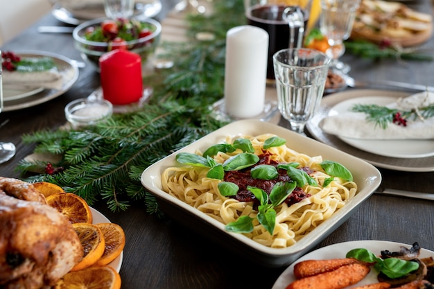 Tavola festiva servita con pasta, tacchino arrosto, piatti, candele, bicchieri da vino, rami di conifere, insalata, dolce fatto in casa e bevande per la cena di Natale