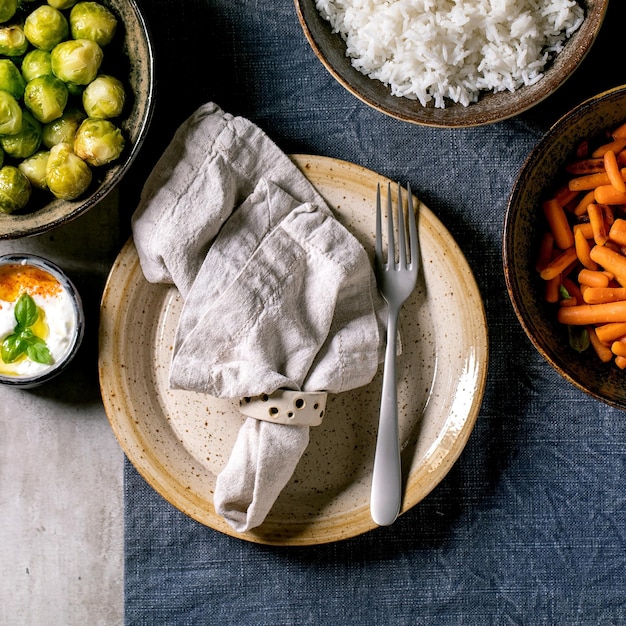 Tavolo da pranzo vegano con riso e verdure cavoletti di Bruxelles al forno carota in ciotole di ceramica e piatto vuoto per gli ospiti su tovaglia di lino blu su sfondo grigio Immagine quadrata piatta