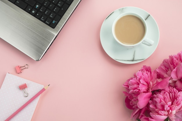 Tavolo da ufficio rosa. Posto di lavoro con laptop, blocco note, fiori di peonia e caffè