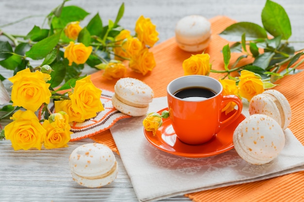 Tazza arancione di caffè nero, fiori di rose gialle e amaretti francesi dolci pastello su tavolo di legno bianco