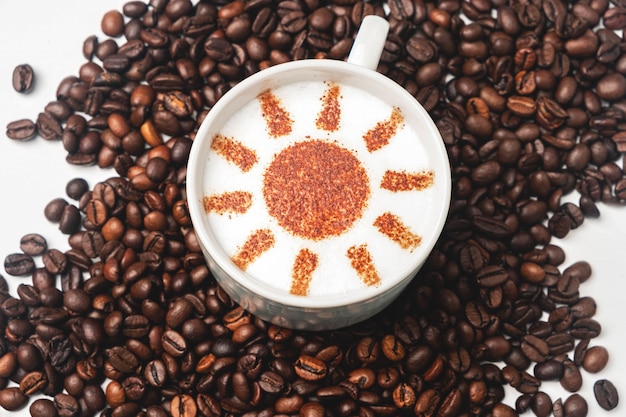 Tazza bianca con caffè cappuccino, motivo sole su schiuma, una superficie di chicchi di caffè.