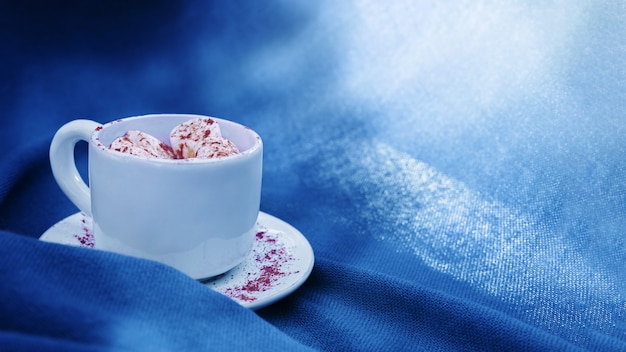 Tazza bianca con caffè e caramelle gommosa e molle alla luce sul fondo del tessuto colorato blu classico con lo spazio della copia. Bevanda calda per la mattina.