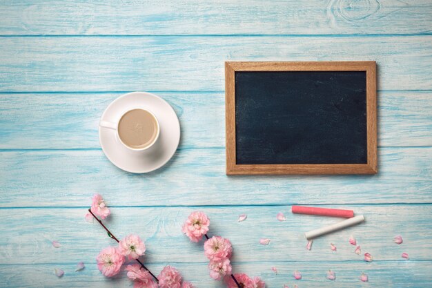 Tazza bianca con cappuccino, fiori di sakura e lavagna su una tavola di legno blu.