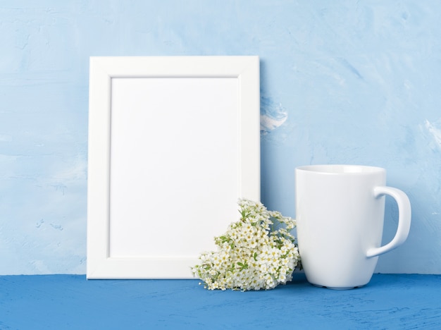 Tazza bianca con tè o caffè, telaio, bouquet di fiori sul tavolo blu di fronte al muro blu.