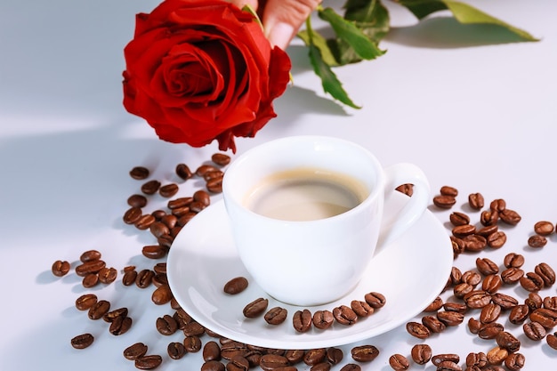 Tazza bianca di caffè nero con una rosa rossa e chicchi di caffè sparsi su un tavolo bianco.